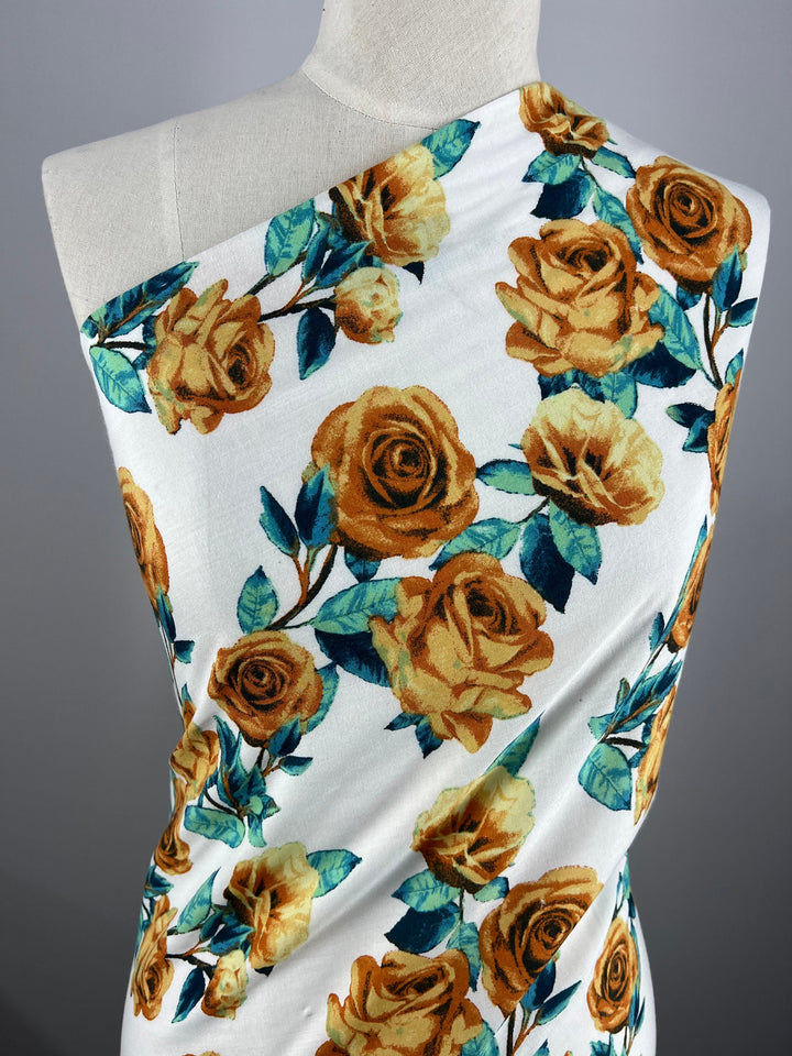 A mannequin draped with **Super Cheap Fabrics' Printed Rayon - La Flora - 145cm**. The lightweight fabric covers one shoulder and wraps diagonally, showcasing the yellow-orange roses and green leaves floral pattern against a gray background.