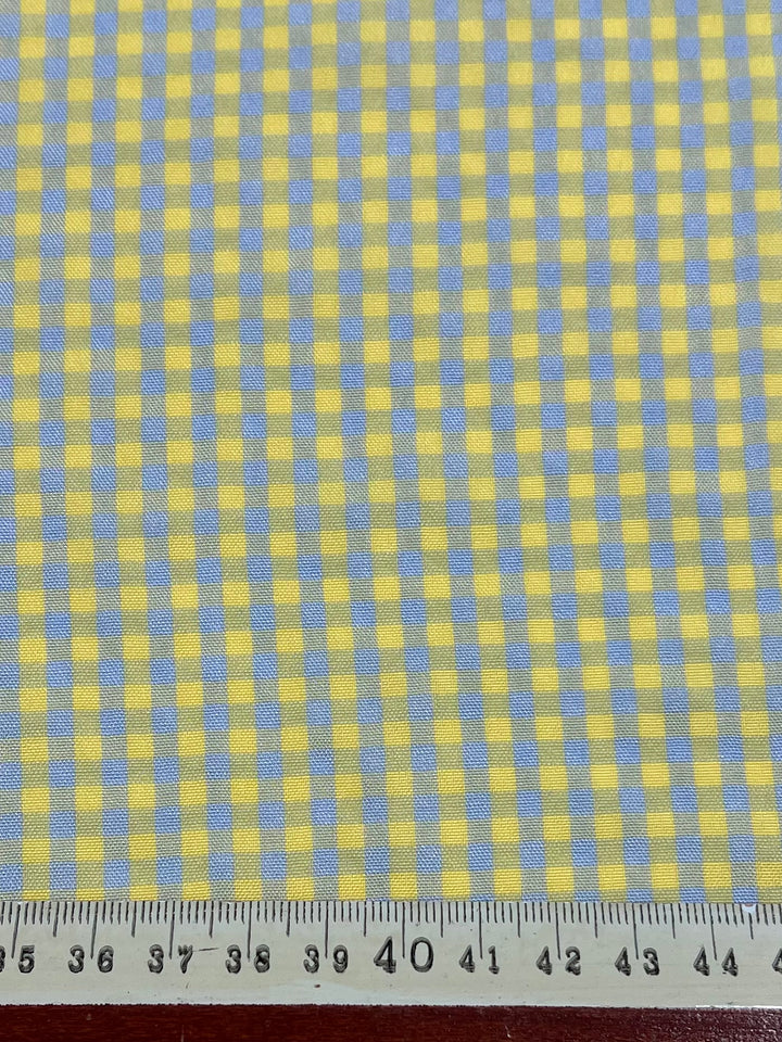 A close-up image of yellow and light blue gingham cotton polyester fabric with a checkered pattern. A ruler placed at the bottom shows measurements in centimeters from 35 to 44, indicating the scale of ***Super Cheap Fabrics' Cotton Poly - Mini Gingham Blue & Yellow- 150cm*** lightweight fabric.