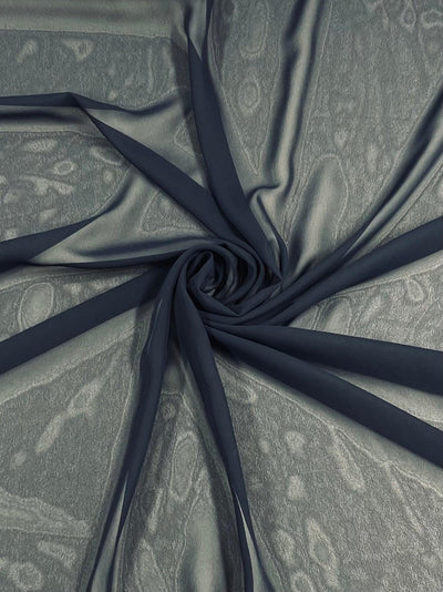 A piece of Hi-Multi Chiffon - Dark Navy - 150cm from Super Cheap Fabrics is arranged in a spiral design on top of a textured, lighter-colored surface. The lightweight fabric appears smooth and delicate, with intricate patterns subtly visible underneath.