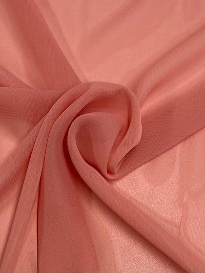 A close-up of a piece of Hi-Multi Chiffon - Coral Almond - 150cm from Super Cheap Fabrics. The lightweight fabric, made from 100% polyester, is slightly gathered and twisted in a gentle swirl at the center, creating subtle folds and texture. The material appears delicate and semi-transparent, with a smooth and silky surface.