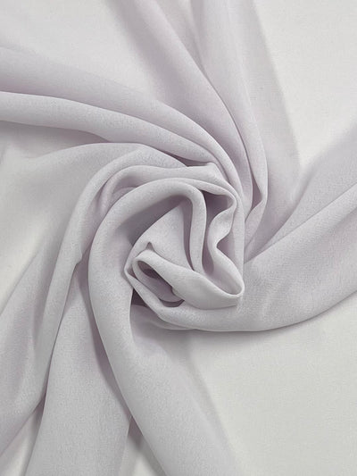 A close-up of sheer, light lavender-colored fabric draped and folded in a circular, swirl-like pattern. The lightweight Georgette texture appears soft and slightly textured, creating gentle shadows and highlights on its surface. This is the "Plain Georgette - Orchid Ice - 112CM" from Super Cheap Fabrics.