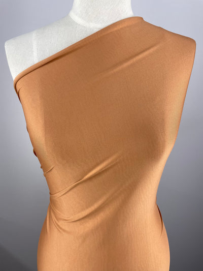A mannequin is draped in a single-shoulder, form-fitting, camel-colored fabric made from medium weight fabric, showcasing its smooth texture and subtle sheen. The Super Cheap Fabrics ITY Knit - Meerkat - 150cm elegantly wraps around the torso, emphasizing its drape and flow. The background is a plain gray.