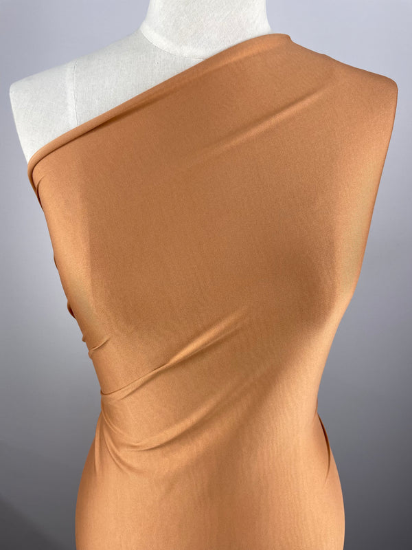 A mannequin is draped in a single-shoulder, form-fitting, camel-colored fabric made from medium weight fabric, showcasing its smooth texture and subtle sheen. The Super Cheap Fabrics ITY Knit - Meerkat - 150cm elegantly wraps around the torso, emphasizing its drape and flow. The background is a plain gray.