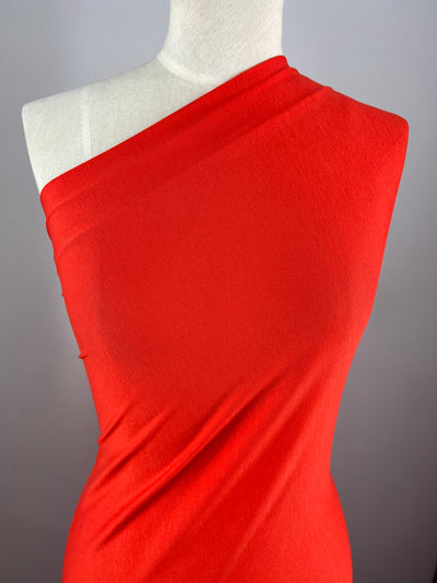 A red, one-shoulder dress displayed on a dress form against a plain grey background. The Rayon Lycra - Red Alert - 150cm from Super Cheap Fabrics has a smooth, sleek texture and the medium weight drapes elegantly over the form, highlighting the contours with its two-way stretch.