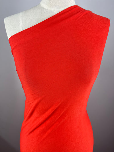 A white mannequin is draped in a single-shouldered, vibrant Rayon Lycra - Coral - 150cm from Super Cheap Fabrics that fits closely to show the contours of the torso. The background is plain gray, putting full focus on the bright, smooth, and elegantly wrapped medium weight fabric.