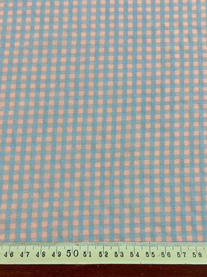 A close-up of a checkered cotton polyester fabric with a pattern of light pink and light blue squares. A ruler placed at the bottom shows measurements in centimeters, with the 50 cm mark centered. The background is a wooden surface. The fabric featured is "Cotton Poly - Mini Gingham Pale Pink & Blue - 150cm" by "Super Cheap Fabrics.