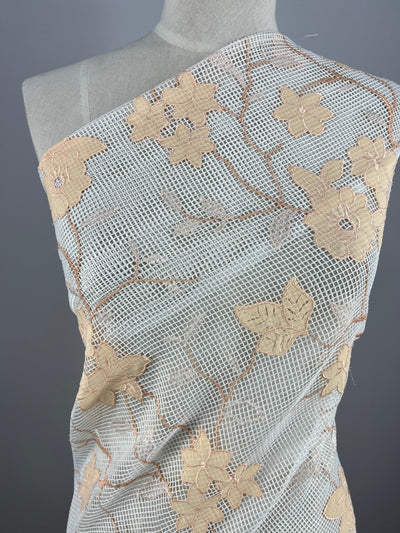 A mannequin draped in Super Cheap Fabrics' Embellished Netting - Apricot with an off-white base and embellished with intricate embroidered floral patterns in shades of beige and gold. The flowers are connected by delicate vines, creating an elegant and decorative design.