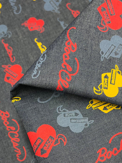 A close-up of folded fabric featuring a fun heart-themed print design. The dark background fabric, made from 100% cotton, showcases colorful patterns, including red, yellow, and blue hearts with "Base" and other text inscriptions, as well as stylized "BadBoi" text in red. Super Cheap Fabrics' Denim - Banner Hearts - 150cm is perfect for a cold wash.