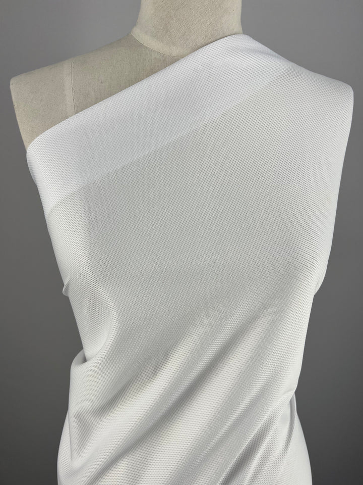 A close-up image of a mannequin draped in a Sportswear Jersey - White -160cm from Super Cheap Fabrics that covers one shoulder, creating an asymmetrical look. The material appears to have a slight sheen and a subtle, small grid pattern, providing generous stretch for maximum comfort. The background is solid gray.