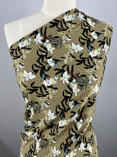 A dress form draped with tan, 100% polyester fabric adorned with a floral pattern. The design features white and red flowers, green foliage, and black accents on a subtle dotted background. The lightweight Designer Georgette - Spot Bouquet by Super Cheap Fabrics is draped to create an asymmetrical look over the shoulder.