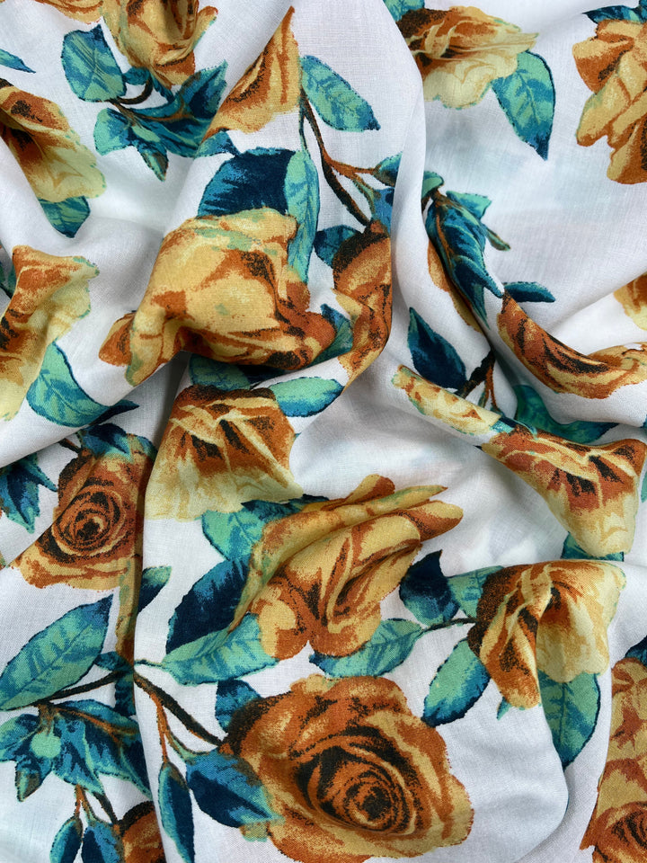 A piece of lightweight fabric with a floral print featuring yellow roses and green leaves on a white background. The 100% rayon material, Printed Rayon - La Flora - 145cm by Super Cheap Fabrics, is slightly wrinkled, showing off the detailed design and vibrant colors.