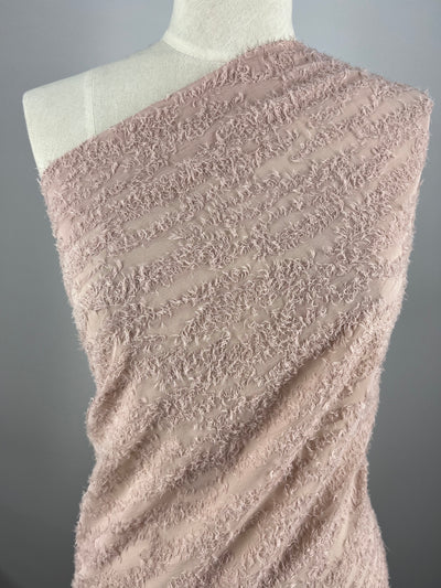 A close-up of a fabric draped on a dress form. The light pink, 100% polyester fabric boasts a textured, fringed pattern, and it covers one shoulder of the dress form, creating a stylish, asymmetrical look. The background is plain and gray, accentuating the Designer - Dusty Pink Texture - 145cm fabric's texture and color from Super Cheap Fabrics.