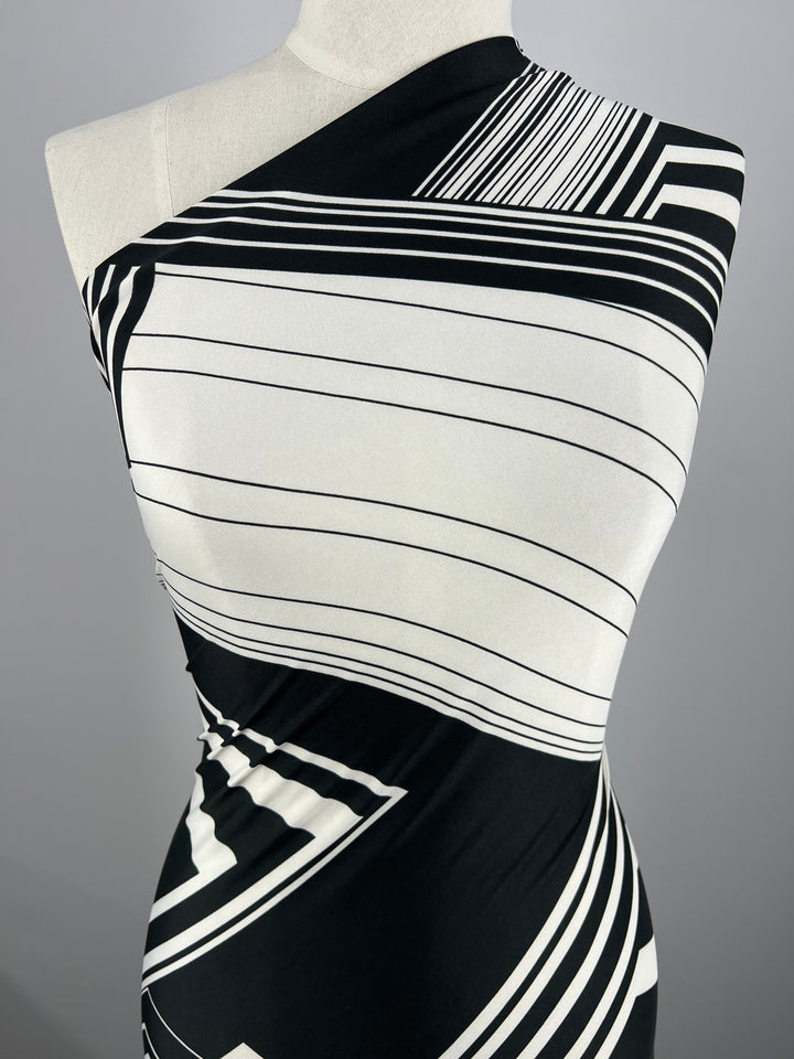 A one-shouldered, close-fitting dress crafted from lightweight Printed Lycra - Diamond Grid by Super Cheap Fabrics with a monochrome geometric design of black and white lines and shapes. The pattern features diagonal and horizontal lines, creating a modern and stylish look. The dress is displayed on a mannequin against a gray background.