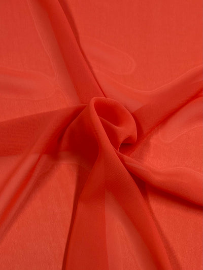 A close-up of a piece of smooth, red fabric. The lightweight fabric is arranged with gentle folds and drapes, creating a soft, flowing appearance. The material is slightly sheer, allowing light to pass through and highlighting its delicate texture. This fabric is Hi-Multi Chiffon - Emberglow - 150cm by Super Cheap Fabrics.