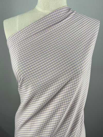 A mannequin is draped with a light-weight fabric featuring a small gingham check pattern in light purple and white. The Cotton Poly - Mini Gingham Peach & Purple - 150cm by Super Cheap Fabrics covers one shoulder, creating a soft, elegant look. The plain gray background highlights the multi-use fabric's exquisite design.