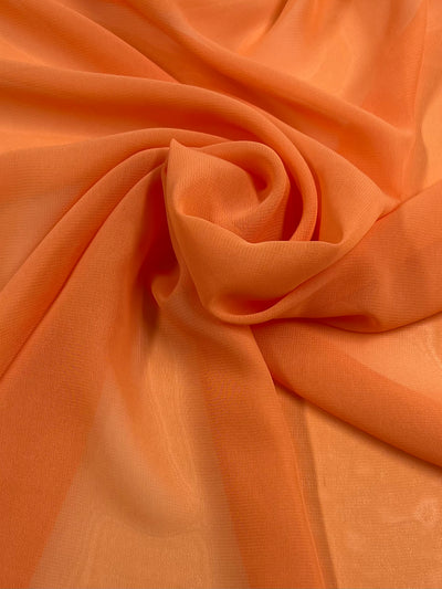 An image of Super Cheap Fabrics Hi-Multi Chiffon - Orange - 150cm, gently gathered and swirled to create folds and textures. The sheer fabric appears smooth, glossy, and lightweight, catching light to showcase varying shades of orange.