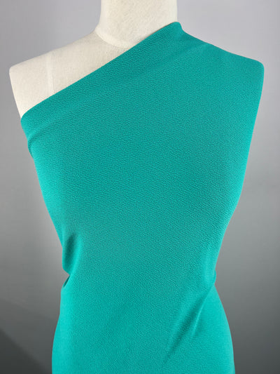 A close-up image of a dress form wearing a vibrant teal, one-shoulder garment made from Super Cheap Fabrics' Textured Knit - Aqua - 150cm. The stretch fabric appears textured and clings smoothly to the form, highlighting a clean and elegant design. The background is a plain, neutral color, making the garment stand out.