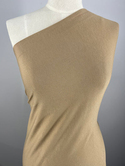 A mannequin displays a tan one-shoulder dress against a grey background. The fabric, Super Cheap Fabrics' Rayon Lycra - Savannah Tan - 150cm, drapes smoothly over the form, showcasing a minimalistic and elegant design.