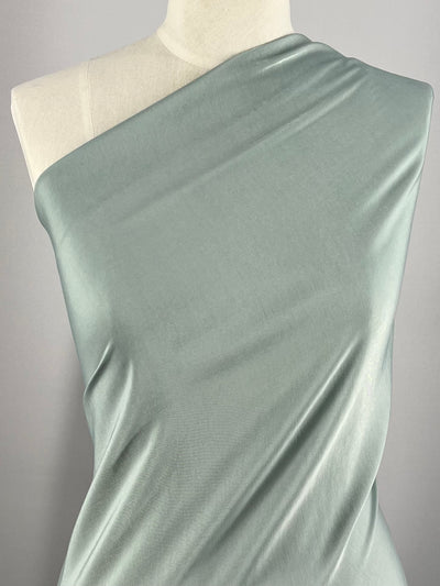 A light green, silky microfiber suiting fabric is draped over a dress form. The lightweight fabric has a smooth texture and shines softly under the light. The dress form is positioned against a plain gray background, showcasing the Microfibre - Gray Mist - 150cm by Super Cheap Fabrics's drape and sheen.