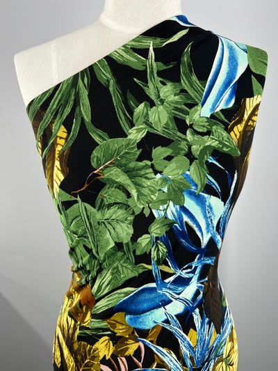 A close-up of a mannequin draped in Super Cheap Fabrics' Printed Lycra - Wilderness Blue - 150cm, featuring a vibrant tropical print on medium weight Polyester/Spandex fabric. The pattern includes green leaves, yellow foliage, and blue elements on a black background, giving a lush and striking appearance.