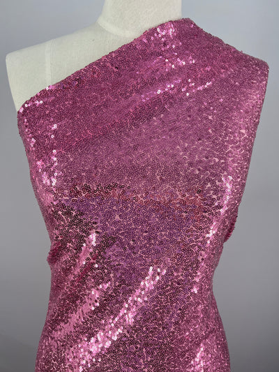 A dress form is draped with a sparkling pink sequin fabric, Evening Sequins - Prism Pink - 150cm by Super Cheap Fabrics, perfect for festival wear. The fabric is arranged in a one-shoulder design, showcasing its shiny and reflective surface. This statement garment stands out beautifully against the plain, neutral background.