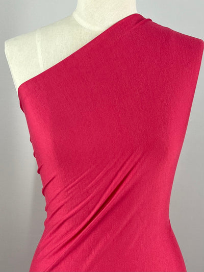A mannequin is draped in a piece of Rayon Lycra - Party Punch - 150cm from Super Cheap Fabrics. The medium weight fabric, fitted snugly, is positioned to show an asymmetrical, one-shoulder design. The background is plain and light gray, allowing the vibrant color of the two-way stretch fabric to stand out.