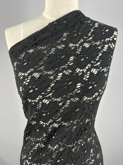A close-up of a one-shoulder black lace dress displayed on a mannequin. The Super Cheap Fabrics Lace - Black - 102cm features an intricate floral pattern, and the dress is form-fitting, highlighting the detailed design of the fabric against a plain grey background.