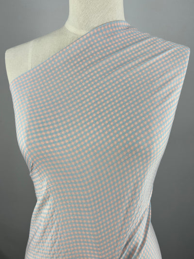A mannequin is dressed in a multi-colour garment with a light blue and pink checkered pattern. The lightweight fabric, composed of Cotton Poly - Mini Gingham Pale Pink & Blue - 150cm by Super Cheap Fabrics, is draped asymmetrically, covering one shoulder and leaving the other bare. The background is a plain gray.