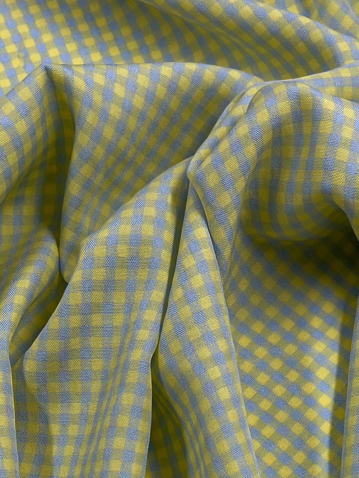 A close-up image of a light weight fabric with a checkered pattern. The pattern consists of alternating small squares in light blue and yellow colors. The Super Cheap Fabrics Cotton Poly - Mini Gingham Blue & Yellow- 150cm fabric has gentle folds, creating soft shadows and highlights.