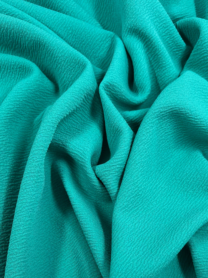 A close-up shot of a vibrant teal, medium weight fabric with a visible textured pattern. The Super Cheap Fabrics Textured Knit - Aqua - 150cm stretch fabric is gently gathered into soft, flowing folds, creating a dynamic interplay of light and shadow on its surface.